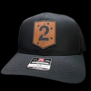 May include: Black trucker hat with a brown leather patch featuring the number 2, a sword, and stars. The hat has a curved brim and a mesh back. The Richardson brand label is visible on the front.