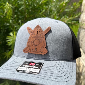 May include: A gray and black trucker hat with a brown leather patch featuring the 1st Marine Division logo. The patch has a star, eagle, globe, and anchor, and the number 8. The hat has a Richardson 112 adjustable strap.