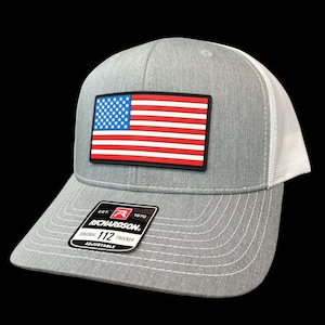 May include: Gray trucker hat with a white mesh back and a rectangular patch featuring the American flag. The hat has a curved brim and the Richardson 112 Trucker label.