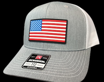 Puede incluir: Gorra de camionero gris con la parte trasera de malla blanca y un parche rectangular con la bandera americana. La gorra tiene una visera curva y la etiqueta Richardson 112 Trucker.