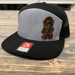 May include: Black and gray flat bill snapback hat with a brown leather patch featuring a cartoon character. The hat has a mesh back and the words "Flat Bill Snapback" are printed on a red tag.