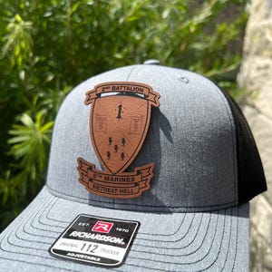 May include: A gray and black trucker hat with a brown leather patch that says "2nd Battalion 1 5th Marines Retreat Hell". The hat is adjustable and has a Richardson logo on the side.