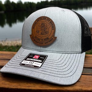 May include: A gray trucker hat with a black mesh back. The hat features a brown leather patch with the text "SECOND BATTALION 4TH MARINES" and "SECOND TO NONE THE MAGNIFICENT BASTARDS". The hat has a curved brim and an adjustable strap.
