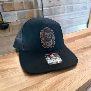 May include: Black trucker hat with a brown leather patch featuring a skull and crossbones with the text "Death Before Decaf". The hat has a black mesh back and a Richardson 112 adjustable strap.