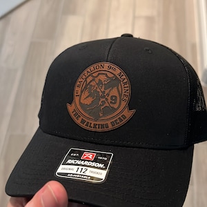 May include: Black trucker hat with a brown leather patch that says "1st Battalion 9th Marines The Walking Dead". The hat is adjustable and has a Richardson logo.