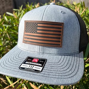May include: A gray trucker hat with a black mesh back and a brown leather American flag patch. The hat has a curved brim and an adjustable strap. The Richardson brand label is visible on the front.