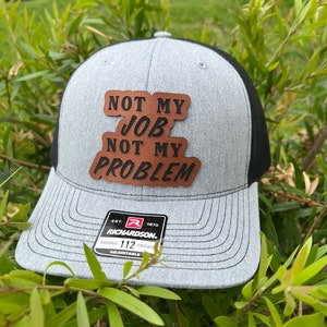 May include: A gray and black trucker hat with a brown leather patch that reads "NOT MY JOB NOT MY PROBLEM". The hat is adjustable and has a Richardson logo on the back.