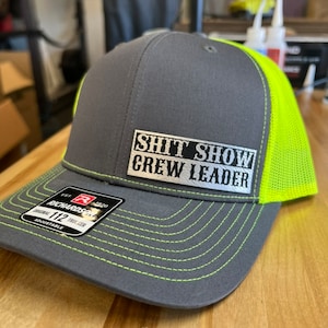 May include: A gray and neon green trucker hat with a black and silver patch that reads "Shit Show Crew Leader".