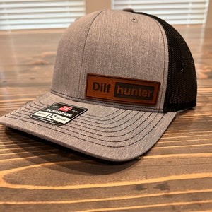 May include: A gray and black trucker hat with a brown leather patch that says "Dilf hunter".