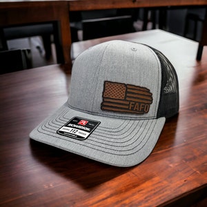 May include: A gray and black trucker hat with a brown leather patch featuring an American flag and the text "FAFO". The hat is adjustable and has a Richardson 112 label.