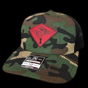 May include: Camouflage trucker hat with a black mesh back and a red emblem featuring an eagle, globe, and anchor. The hat has a curved brim and a Richardson tag. The hat is adjustable.