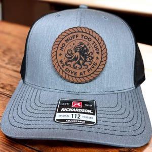 May include: A gray and black trucker hat with a brown leather patch featuring an octopus and the text "No Muff Too Tuff We Dive At Five". The hat is adjustable and has the Richardson logo and the text "Original 112 Trucker".