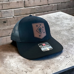 May include: A black and brown trucker hat with a leather patch featuring a skull and stars. The patch says "1st 1 1st". The hat is made by Richardson.
