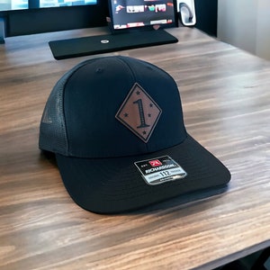 May include: A black and brown trucker hat with a brown leather patch featuring a diamond shape with a white number "1" and stars. The hat has a black mesh back and a Richardson label with the text "Original Trucker 112 Adjustable".