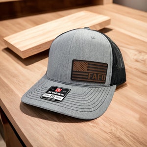 May include: A gray and black trucker hat with a brown leather patch featuring a US flag and the text "FAFO". The hat has a Richardson 112 Original Trucker label.