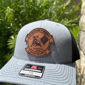 May include: Gray and black trucker hat with a leather patch featuring a bull and text that reads "3rd Battalion 1st Marines, Thundering Third, Balls of the Corps." The hat has a curved brim and an adjustable strap.