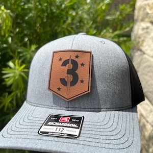 May include: A gray and black trucker hat with a brown leather patch featuring a stylized number "3" and a dagger. The patch has four stars around the number. The hat has a black mesh back and a label that reads "EST. 1970 RICHARDSON ORIGINAL 112 TRUCKER ADJUSTABLE".