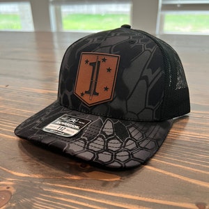 May include: A black and gray camouflage trucker hat with a brown leather patch featuring a sword and the number "1" surrounded by stars. The hat is adjustable and has a mesh back.