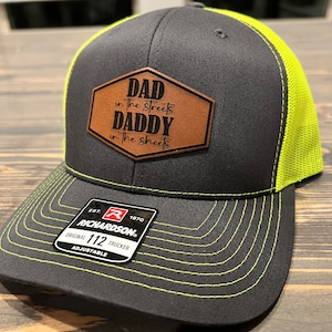 May include: A black and neon green trucker hat with a brown leather patch that reads "DAD in the streets DADDY in the sheets". The hat is adjustable and has a Richardson 112 logo.