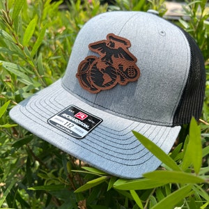 May include: A gray and black trucker hat with a brown leather patch featuring the United States Marine Corps emblem. The patch is stitched onto the front of the hat. The hat is adjustable and has a Richardson 112 label.