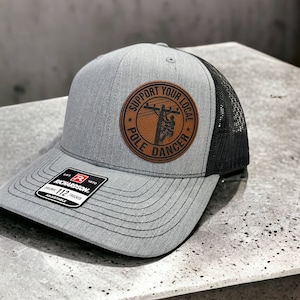 May include: A gray and black trucker hat with a brown leather patch that says "Support Your Local Pole Dancer".