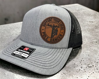 May include: A gray and black trucker hat with a brown leather patch that says "Support Your Local Pole Dancer".