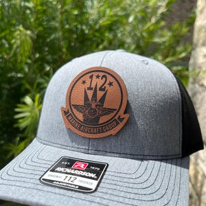 May include: A gray and black trucker hat with a brown leather patch featuring a winged emblem and the text "*1*2* MARINE AIRCRAFT GROUP". The hat also has a red and black label with the text "EST. 1970 RICHARDSON. ORIGINAL 112 TRUCKER ADJUSTABLE".