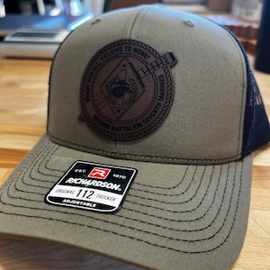 May include: A green and black baseball cap with a brown leather patch that reads "READY FOR ALL YIELDING TO NONE * SECOND BATTALION SEVENTH MARINES". The cap is a trucker style with a mesh back and a Richardson 112 label.