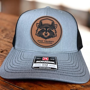 May include: A gray and black trucker hat with a brown leather patch featuring a raccoon wearing a hat and the text "Stay Trashy". The hat has a black mesh back and an adjustable strap with the Richardson 112 logo.