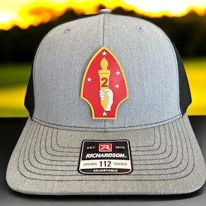 May include: Gray and black trucker hat with a patch featuring a torch, the number 2, and stars on a red background. The hat has a Richardson 112 Trucker label.