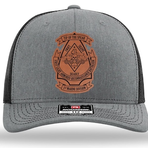 May include: Gray trucker hat with a black mesh back and a curved brim. The front features a brown patch with the text "TIP OF THE SPEAR", "SWIFT", "SILENT", and "DEADLY". The patch also includes the text "FIRST LIGHT ARMORED RECONNAISSANCE BATTALION" and "1ST MARINE DIVISION".