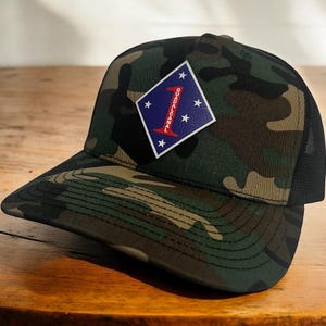 May include: A camouflage trucker hat with a black mesh back. The hat features a black diamond patch with a red "1" and the text "GUADALCANAL" in white.