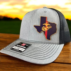 May include: A gray and black trucker hat with a leather patch featuring the outline of the state of Texas with the Marine Corps emblem and a red stripe down the center. The hat has the text "Richardson 112 Trucker Adjustable" on the back.