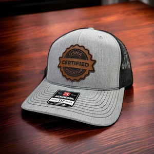 May include: A gray and black trucker hat with a brown leather patch that says "OSHA Certified Violator". The hat is adjustable and has a Richardson 112 label.