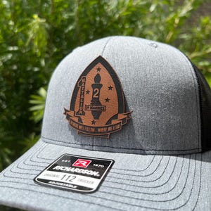 May include: Gray trucker hat with a black mesh back and a leather patch. The patch features the text "20 MARINES" and "OTHERS WILL FOLLOW WHERE WE LEAD". The hat also has a Richardson 112 Trucker tag.