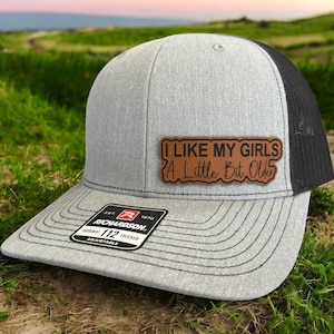 May include: Gray trucker hat with a black mesh back and a curved brim. The hat features a brown leather patch with the text "I LIKE MY GIRLS A Little Bit Older". The hat has a Richardson 112 Trucker tag.