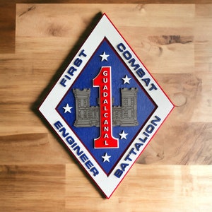 May include: A diamond-shaped emblem with the text "FIRST COMBAT ENGINEER BATTALION GUADALCANAL" in blue lettering. The emblem features a red number 1, two gray castle illustrations, and white stars, set against a blue background.