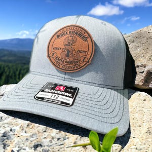 May include: A gray and black trucker hat with a brown leather patch that reads "1st Battalion Ridge Runners First to Fight 1 Baker Company 7th Marines". The hat has a black mesh back and a Richardson 112 adjustable strap.