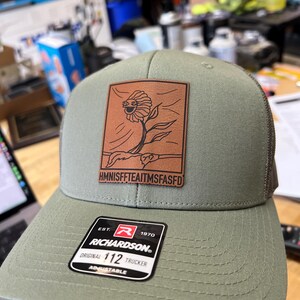 May include: Olive green Richardson 112 trucker hat with a brown leather patch featuring a whimsical flower design. The patch has the text "HMNISFFTEAITMSFASFD". The hat has a mesh back and an adjustable strap.