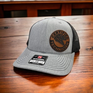 May include: A gray and black trucker hat with a brown leather patch that says "Certified Zip Tie Technician". The hat is adjustable and has a Richardson 112 label.
