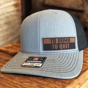 May include: Gray trucker hat with a black mesh back and a curved brim. The hat features a rectangular patch with the words "TO DUMB TO QUIT" in white lettering. The Richardson brand label is visible.