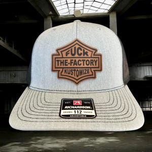 May include: A gray trucker hat with a black mesh back and a brown leather patch. The patch features the words "FUCK THE-FACTORY KUSTOMIZE". The hat has a curved brim and an adjustable strap.
