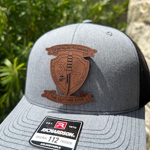 May include: A gray and black trucker hat with a brown leather patch that says "30 Battalion 7th Marines, The Cutting Edge". The patch has a sword and a shield with the number "3" on it. The hat also has a black mesh back and a Richardson logo.