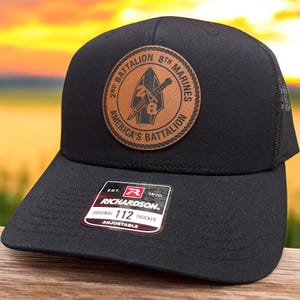 May include: Black trucker hat with a brown leather patch featuring the text "2nd Battalion 8th Marines America's Battalion." The hat has a curved brim and mesh back. The Richardson label reads "Original 112 Trucker Adjustable."
