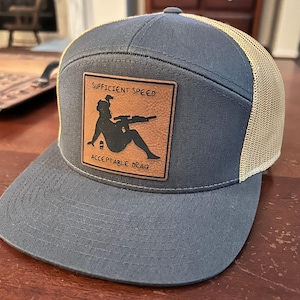 May include: A blue and tan trucker hat with a brown leather patch that says "Sufficient Speed Acceptable Drag" and features a silhouette of a woman holding a rifle.