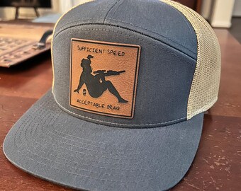 May include: A blue and tan trucker hat with a brown leather patch that says "Sufficient Speed Acceptable Drag" and features a silhouette of a woman holding a rifle.