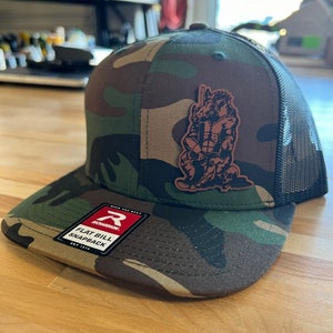 May include: A camouflage trucker hat with a brown leather patch featuring a cartoon rhino wearing a baseball uniform. The hat has a black mesh back and a red "Richardson" label with the text "Flat Bill Snapback".
