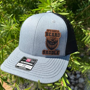 May include: A gray and black trucker hat with a brown leather patch that says "It's not a beard it's a saddle".