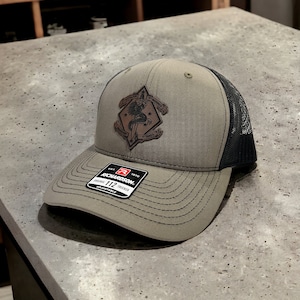 May include: A green and black trucker hat with a brown leather patch featuring a skull and crossbones design. The patch has the text "1ST BATTALION" and "MARINE" on the top and "IT TAKES" on the bottom. The hat has a Richardson 112 adjustable trucker label.