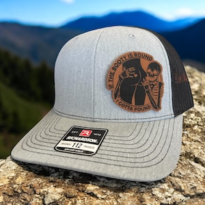 May include: Gray trucker hat with a black mesh back and a curved brim. The hat features a brown leather patch with the text "IF THE BOOTY IS ROUND I GOTTA POUND" and a graphic of a skeleton and a butt. The hat has the Richardson logo.